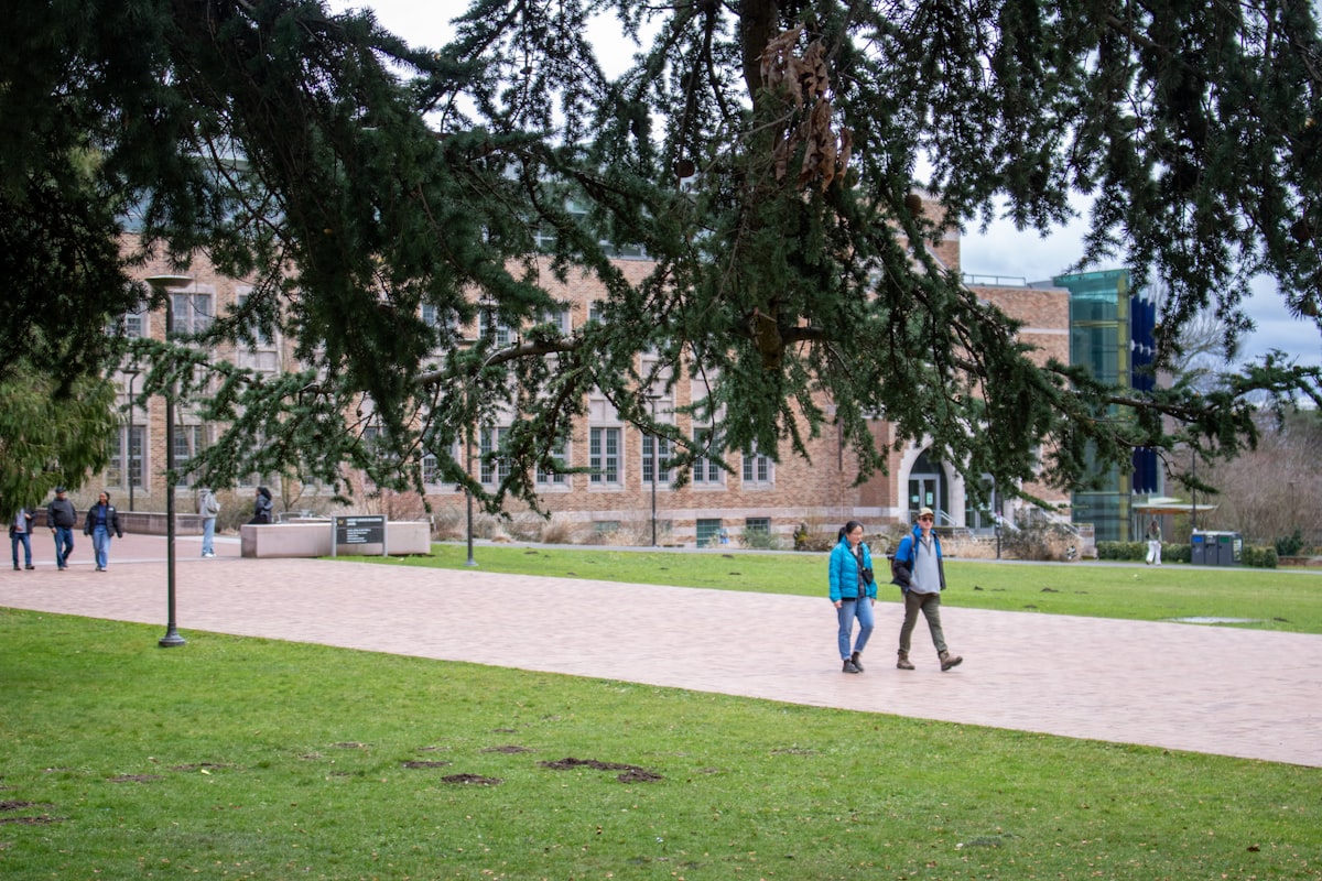 Zwei Studierende gehen unter herabhängenden Baumzweigen über den herbstlichen Campus vor einem Backsteingebäude als Symbol für den Semesterstart und den Empfang neuer Erstsemester