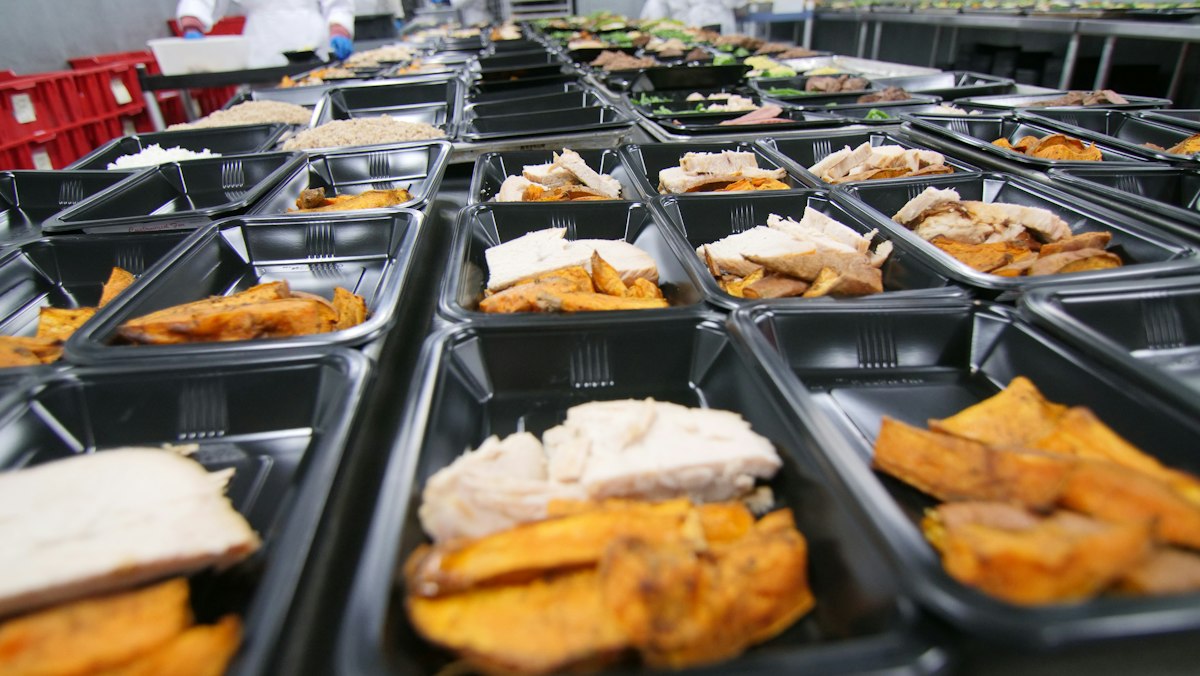 Reihen portionierter Mittagessen mit Hähnchen und Süßkartoffeln in Menüschalen in einer Großküche, Symbol für die Produktion in der Schul- und Kitaverpflegung